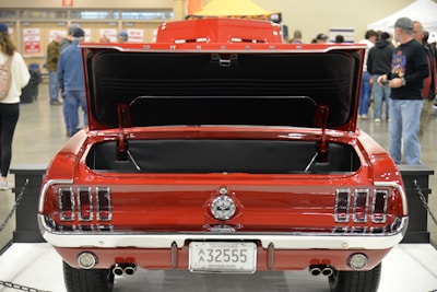 A classic red car with its trunk open is displayed indoors. Several people are visible in the background, suggesting a car show or exhibition setting. The car's license plate indicates it is an antique vehicle from Tennessee. The setting appears busy with various attendees walking around the venue.