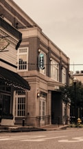 A street view featuring a brick building housing a bank, with large windows and an archway entrance. The scene includes a nearby older-style storefront and a visible part of the street with painted lines. There are streetlights and greenery in the form of trees and planters.