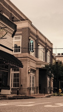 A street view featuring a brick building housing a bank, with large windows and an archway entrance. The scene includes a nearby older-style storefront and a visible part of the street with painted lines. There are streetlights and greenery in the form of trees and planters.