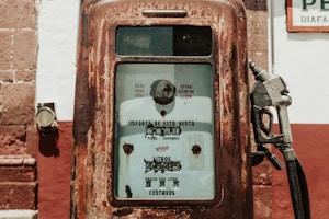 An old, rusted gas pump with visible text in Spanish indicating the amount of fuel dispensed and price. It is positioned against a wall with contrasting red and white colors and a section of a brown brick structure.