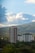 Tall buildings rise among lush greenery, set against a backdrop of rolling hills and a sky filled with large, fluffy clouds. The scene conveys a balance between urban development and natural landscape.