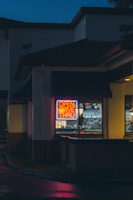 A neon sign reading 'Open Drive Thru' glows brightly in the window of a building at night. The scene captures the exterior corner of the structure, with dark surroundings and a hint of light reflecting off wet pavement. The atmosphere is quiet and somewhat subdued, with the vibrant colors of the sign contrasting against the dimly lit environment.