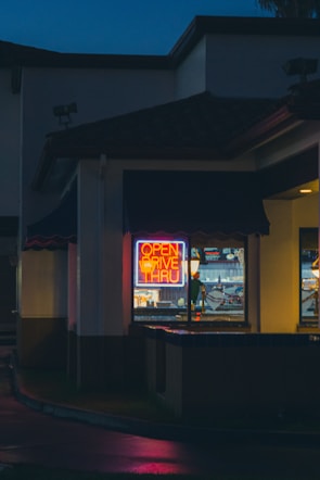 A neon sign reading 'Open Drive Thru' glows brightly in the window of a building at night. The scene captures the exterior corner of the structure, with dark surroundings and a hint of light reflecting off wet pavement. The atmosphere is quiet and somewhat subdued, with the vibrant colors of the sign contrasting against the dimly lit environment.