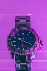 A close-up of a luxury watch with a metallic strap, featuring a blue face and silver detailing. The watch is illuminated with a vibrant purple light that adds a modern and stylish aesthetic.