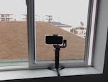 A smartphone is mounted on a gimbal or tripod placed on a windowsill, with a view of rooftops, residential buildings, and a single palm tree visible through the window.