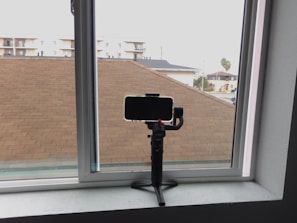 A smartphone is mounted on a gimbal or tripod placed on a windowsill, with a view of rooftops, residential buildings, and a single palm tree visible through the window.