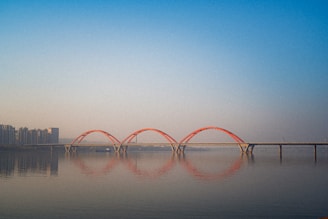 A modern bridge with three red arch supports spans across a calm body of water, reflecting its shape on the water's surface. The skyline features a row of high-rise buildings on the left, fading into the distance under a clear blue sky.