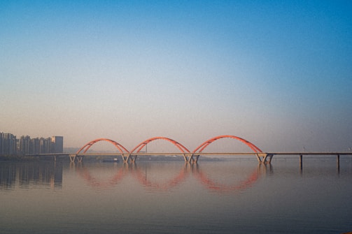 A modern bridge with three red arch supports spans across a calm body of water, reflecting its shape on the water's surface. The skyline features a row of high-rise buildings on the left, fading into the distance under a clear blue sky.