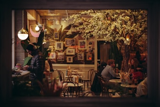 A cozy cafe scene with several people seated at tables, engaging in conversation. A large decorative tree with white blossoms adds a touch of natural elegance to the warmly lit setting. Framed pictures adorn the walls, and a man in a jacket and headphones is standing at a counter, engrossed in a task.