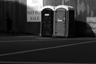Two portable toilets are positioned side by side in front of a corrugated metal wall. A large sign nearby reads 'This Property is NOT FOR SALE', and a person walks away from the scene, partially in shadow.