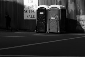 Two portable toilets are positioned side by side in front of a corrugated metal wall. A large sign nearby reads 'This Property is NOT FOR SALE', and a person walks away from the scene, partially in shadow.