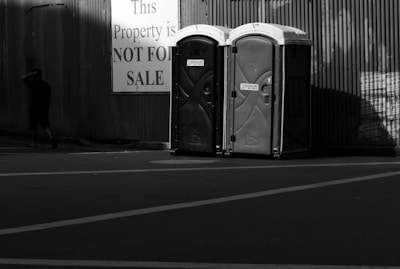 Two portable toilets are positioned side by side in front of a corrugated metal wall. A large sign nearby reads 'This Property is NOT FOR SALE', and a person walks away from the scene, partially in shadow.