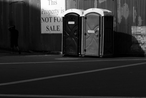 Two portable toilets are positioned side by side in front of a corrugated metal wall. A large sign nearby reads 'This Property is NOT FOR SALE', and a person walks away from the scene, partially in shadow.