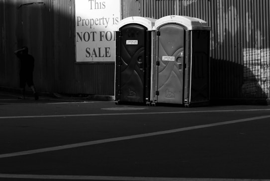 Two portable toilets are positioned side by side in front of a corrugated metal wall. A large sign nearby reads 'This Property is NOT FOR SALE', and a person walks away from the scene, partially in shadow.