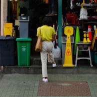 A person with a yellow shirt and beige pants is walking up steps towards a hardware or supply store. The entrance is cluttered with various colorful items such as bins, signs, boots, and cleaning supplies. A 'Caution Wet Floor' sign is prominently placed near the entrance.