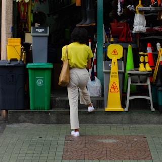 A person with a yellow shirt and beige pants is walking up steps towards a hardware or supply store. The entrance is cluttered with various colorful items such as bins, signs, boots, and cleaning supplies. A 'Caution Wet Floor' sign is prominently placed near the entrance.