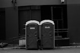 Two portable toilets are placed side by side on an urban street. A small child is partially visible, seemingly trying to open one of the doors. The background consists of a building with dark, closed shutters and an industrial look.