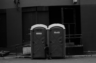 Two portable toilets are placed side by side on an urban street. A small child is partially visible, seemingly trying to open one of the doors. The background consists of a building with dark, closed shutters and an industrial look.