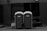 Two portable toilets are placed side by side on an urban street. A small child is partially visible, seemingly trying to open one of the doors. The background consists of a building with dark, closed shutters and an industrial look.
