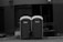 Two portable toilets are placed side by side on an urban street. A small child is partially visible, seemingly trying to open one of the doors. The background consists of a building with dark, closed shutters and an industrial look.