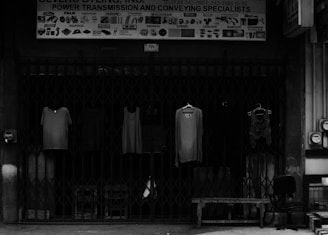 Four pieces of clothing are hung on hangers in front of a closed metal gate. Above the gate is a sign that reads 'Power Transmission and Conveying Specialists.' Various industrial graphics and names are displayed on the sign. To the side, there is an office chair and a bench.