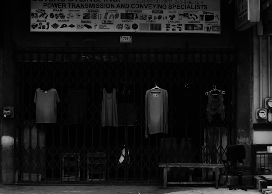 Four pieces of clothing are hung on hangers in front of a closed metal gate. Above the gate is a sign that reads 'Power Transmission and Conveying Specialists.' Various industrial graphics and names are displayed on the sign. To the side, there is an office chair and a bench.