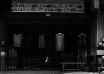 Four pieces of clothing are hung on hangers in front of a closed metal gate. Above the gate is a sign that reads 'Power Transmission and Conveying Specialists.' Various industrial graphics and names are displayed on the sign. To the side, there is an office chair and a bench.