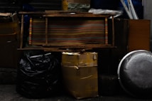 Various objects are stacked against a dark background. There are several large black and yellow garbage bags, a striped wooden bench, a metal pot lid, a cardboard box secured with tape, and other miscellaneous items.