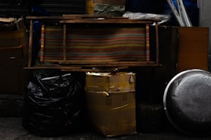 Various objects are stacked against a dark background. There are several large black and yellow garbage bags, a striped wooden bench, a metal pot lid, a cardboard box secured with tape, and other miscellaneous items.