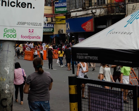 A bustling urban street scene with various people walking and congregating near orange barricades. Signs for Grab Food and foodpanda are visible on a post, and a large tent with the Converge branding is present. The background features several shops, including signs for Magdalena Pawnshop and other local businesses.