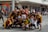 A group of people poses on a street, many wearing coordinated maroon and yellow sports attire. Some individuals are making hand gestures, and a few are wearing face masks. The background features a commercial building with visible signage, and there are other people in the distance.