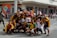A group of people poses on a street, many wearing coordinated maroon and yellow sports attire. Some individuals are making hand gestures, and a few are wearing face masks. The background features a commercial building with visible signage, and there are other people in the distance.