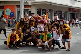 A group of people poses on a street, many wearing coordinated maroon and yellow sports attire. Some individuals are making hand gestures, and a few are wearing face masks. The background features a commercial building with visible signage, and there are other people in the distance.