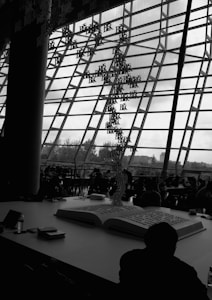 A large open book is placed on a table inside a modern building with a glass-paneled facade. Hanging from the ceiling are alphabetic characters forming an artistic installation. In the background, several people are sitting and working or conversing at tables, partially silhouetted against the windows.