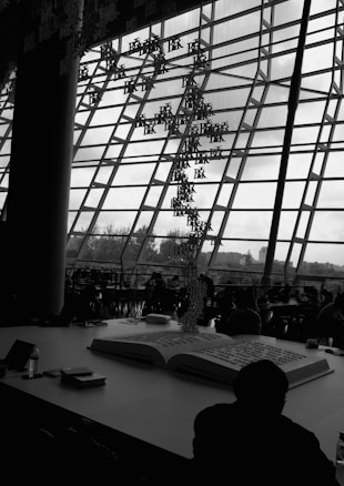A large open book is placed on a table inside a modern building with a glass-paneled facade. Hanging from the ceiling are alphabetic characters forming an artistic installation. In the background, several people are sitting and working or conversing at tables, partially silhouetted against the windows.