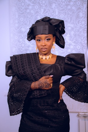 A woman is dressed in elegant traditional attire featuring a black headwrap and a matching intricately designed gown with puffed sleeves. She wears gold jewelry including a necklace and earrings, and her left hand displays detailed henna designs. The background has a subtle white floral pattern.