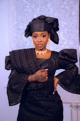A woman is dressed in elegant traditional attire featuring a black headwrap and a matching intricately designed gown with puffed sleeves. She wears gold jewelry including a necklace and earrings, and her left hand displays detailed henna designs. The background has a subtle white floral pattern.
