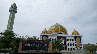 A large mosque with a prominent dome featuring intricate geometric patterns in yellow and green. The mosque has smaller domes and a tall minaret with similar designs. Surrounding the building are trees and greenery, with a signboard in the foreground.
