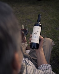 A person is holding a bottle of red wine labeled 'Costa y Roca' and a glass of red wine in their hand while sitting on a chair. The scene is outdoors with grass visible in the background. The person is casually dressed in a plaid shirt and khaki pants.