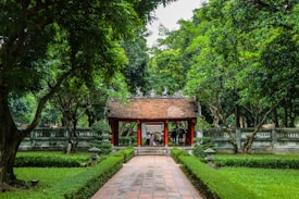 A traditional pavilion with a tiled roof stands at the end of a pathway, flanked by lush green trees and manicured hedges. The scene is tranquil and surrounded by nature, with people visible beneath the structure.