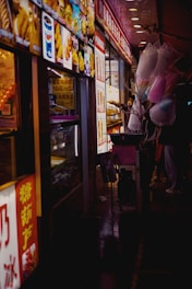 Colorful food stall at night with bright signage and vibrant images of snacks and beverages. Cotton candy in pastel shades of pink and blue is held by a vendor. The scene conveys the bustling atmosphere of a night market.