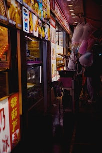 Colorful food stall at night with bright signage and vibrant images of snacks and beverages. Cotton candy in pastel shades of pink and blue is held by a vendor. The scene conveys the bustling atmosphere of a night market.