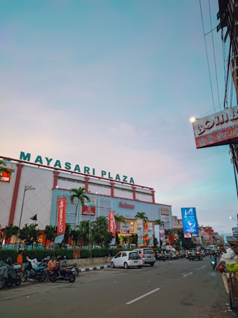 A large shopping mall named 'Mayasari Plaza' with a variety of signage and advertisements on its facade. The building is modern with red and white color themes and has palm trees and banners in front. There are numerous vehicles and motorcycles parked along the street as well as a few people around. The sky is clear with a slight gradient from light to darker blue.