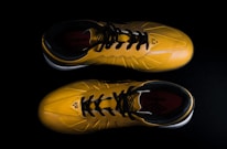 A pair of yellow sports shoes with black laces is placed against a dark background. The design is sleek, with angular patterns on the surface. The inside of the shoes appears to have a red lining with a brand logo visible.