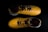A pair of yellow sports shoes with black laces is placed against a dark background. The design is sleek, with angular patterns on the surface. The inside of the shoes appears to have a red lining with a brand logo visible.