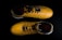 A pair of yellow sports shoes with black laces is placed against a dark background. The design is sleek, with angular patterns on the surface. The inside of the shoes appears to have a red lining with a brand logo visible.