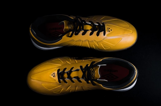A pair of yellow sports shoes with black laces is placed against a dark background. The design is sleek, with angular patterns on the surface. The inside of the shoes appears to have a red lining with a brand logo visible.
