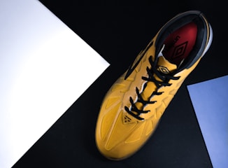 A yellow sports shoe with black laces and a design logo is placed against a black background. The shoe is angled diagonally from bottom left to top right. There are two sheets of paper, one white and one blue, positioned near the edges of the image.