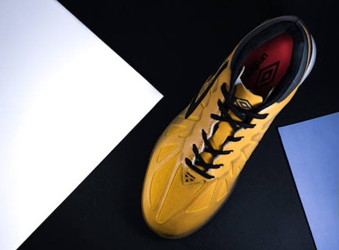 A yellow sports shoe with black laces and a design logo is placed against a black background. The shoe is angled diagonally from bottom left to top right. There are two sheets of paper, one white and one blue, positioned near the edges of the image.