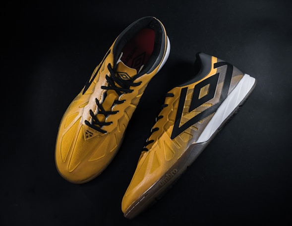 A pair of yellow and black sports shoes displayed against a dark background. The shoes feature a bold geometric pattern and are designed for athletic performance.